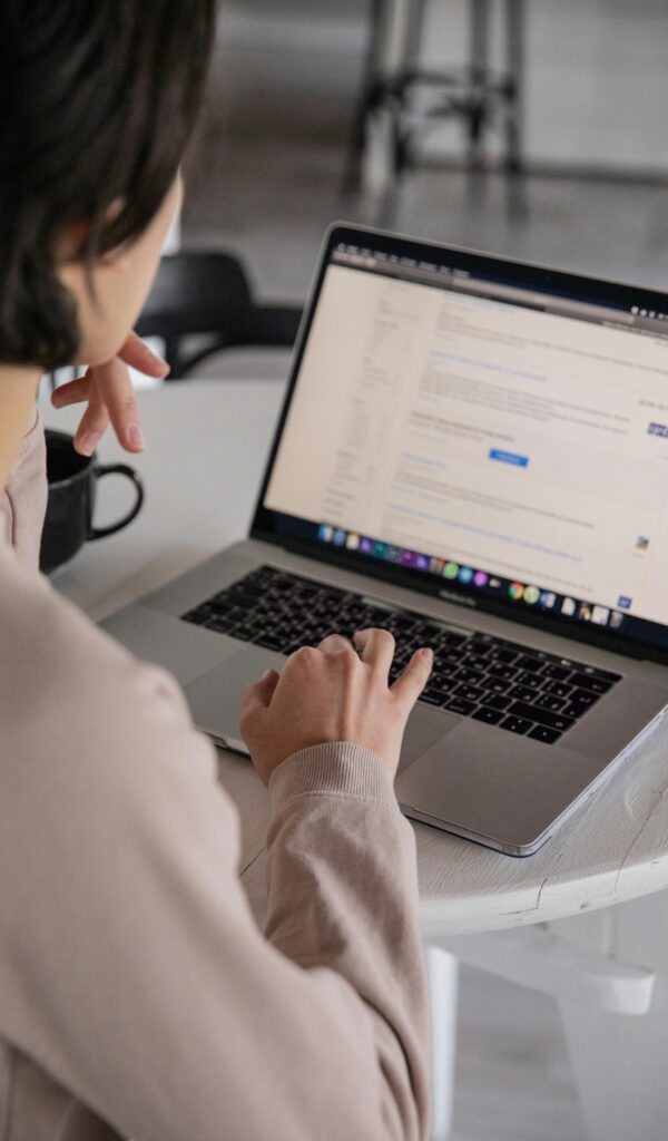 Back view of crop anonymous female remote employee typing on portable computer while watching website on screen and sitting at plastic table with cup of hot drink in apartment